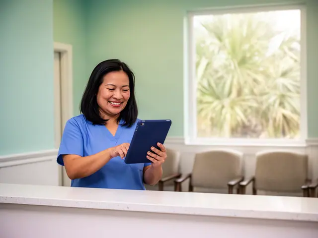 A smiling woman in ceil-blue scrubs using a navy-cased tablet at a dental reception counter.